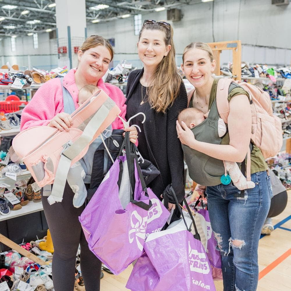 Three ladies smiling with purple JBF shopping bags smiling. One woman is carrying a baby in a front-carrier.