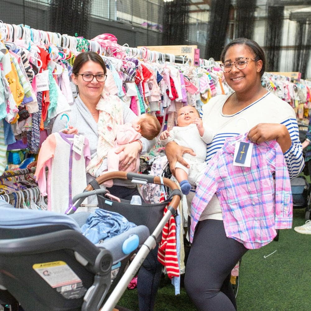 Two moms holding their babies hold up hangers with clothing. They are standing in front of clothing racks.